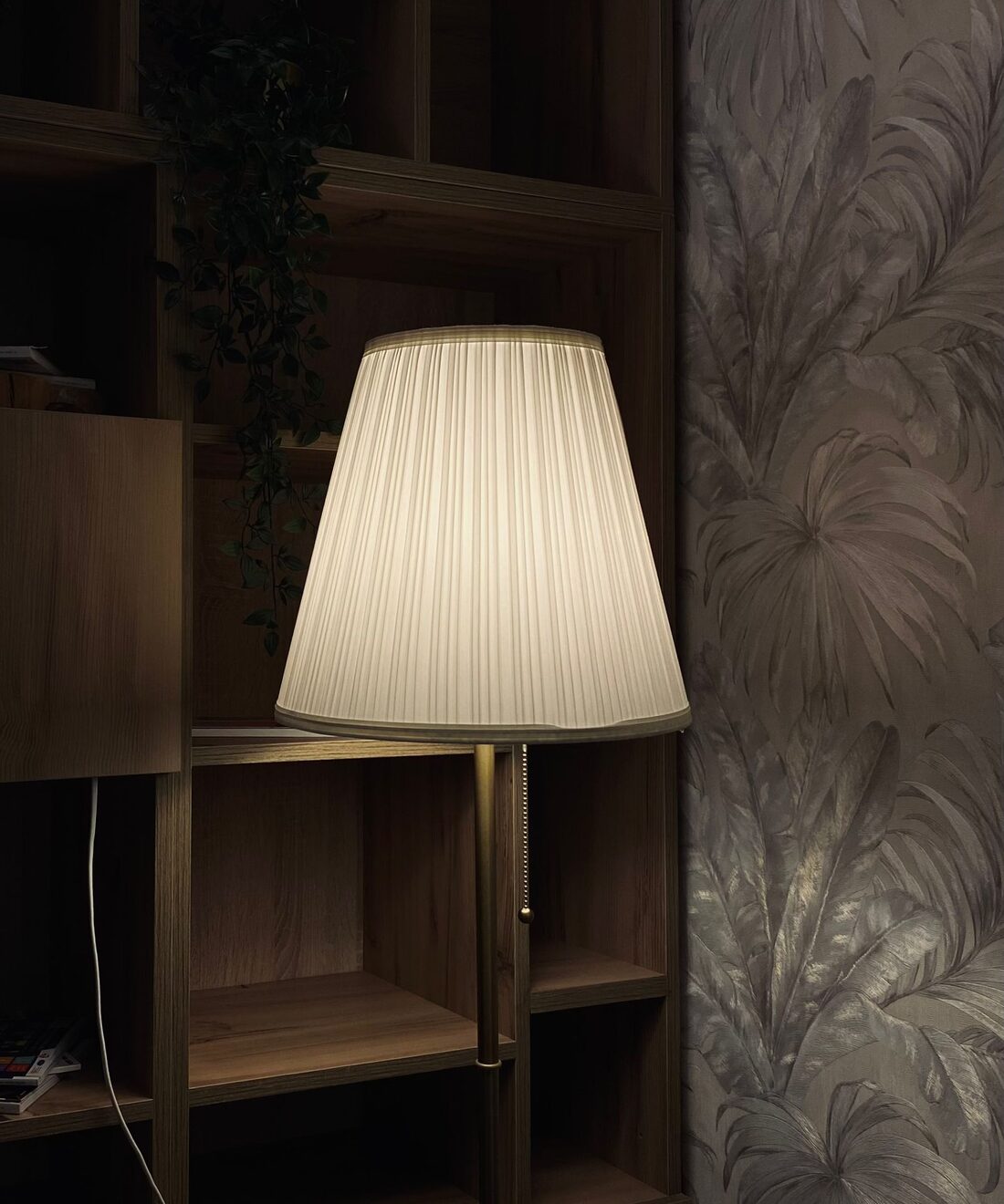 Dark botanical wallpaper feature wall softly lit by a floor lamp beside built-in shelving